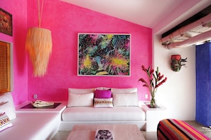 Ein Zimmer mit einer leuchtend rosafarbenen Wand, einem weißen Sofa mit bunten Kissen, einer Pendelleuchte und einem Gemälde über dem Sofa.