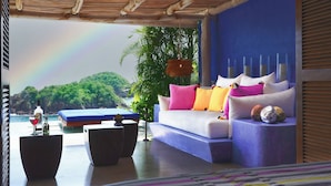 A modern outdoor seating area with a sofa, cushions, and a table, overlooking a lush landscape and a rainbow. - Casita Jazmin Careyes - Beachfront Villa - Infinity Pool - Exoticdigs Collection (Careyes)