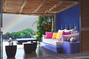 Ein moderner Außenbereich zum Sitzen mit einer Couch, Kissen und einem Tisch, mit Blick auf eine üppige Landschaft und einen Regenbogen.