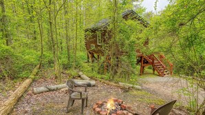 A rustic cabin surrounded by dense forest, with a fire pit and a bench in the foreground.