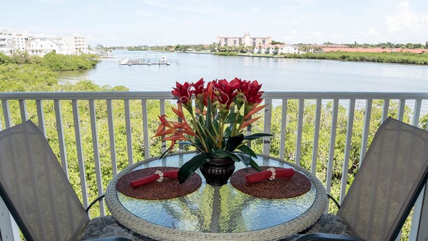 A glass table with two chairs on a balcony overlooking a body of water with buildings in the background. - 140+ Reviews, Steps to Beach, Magnificent Water Views, None Cleaner, None Nicer! (Indian Shores)