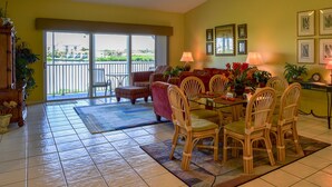 A spacious living area with a dining table surrounded by wicker chairs, a sofa, and a balcony with a view of a residential area. - 140+ Reviews, Steps to Beach, Magnificent Water Views, None Cleaner, None Nicer! (Indian Shores)