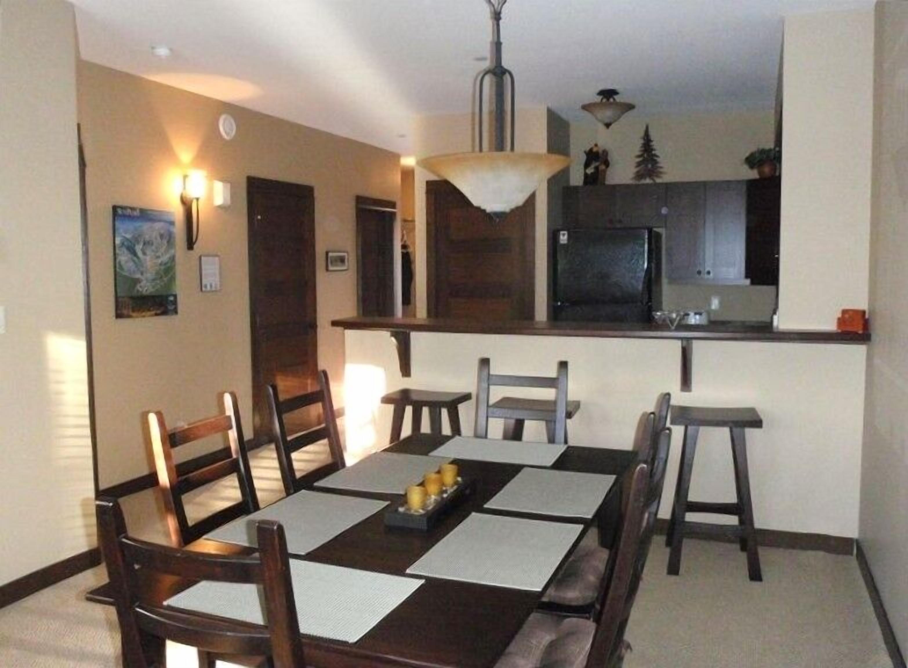 Comedor con mesa preparada para cuatro, zona de cocina con refrigerador y microondas, y lámpara colgante.