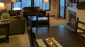 Una sala de estar acogedora con chimenea, un sofá, una mesa de centro y un televisor. Hay dos ventanas con cortinas, y una lámpara está a la izquierda.