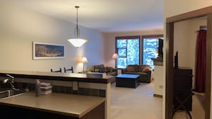 Una cocina moderna con una isla central, un comedor con mesa y sillas, una sala de estar con sofá y una mesa de centro, y un ventanal con vistas a árboles.
