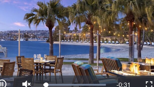 Un área de comedor frente a la playa con mesas y sillas, palmeras y vistas al mar y al paisaje urbano.