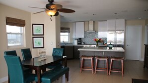 Una cocina moderna con una isla central, mesa de comedor de madera oscura y sillas color turquesa, y un ventilador de techo.