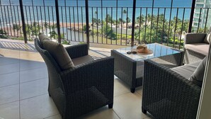 Un balcón con mobiliario de mimbre y una mesa de vidrio, con vista a una playa y al océano.
