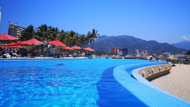 Una piscina azul cristalina con borde curvo, sombrillas rojas y palmeras.