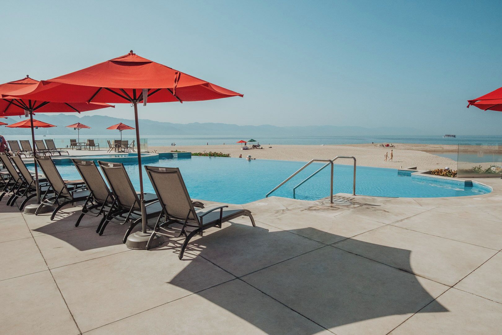 Zona de piscina con sillones de descanso y sombrillas rojas, con vista a una playa y al océano.