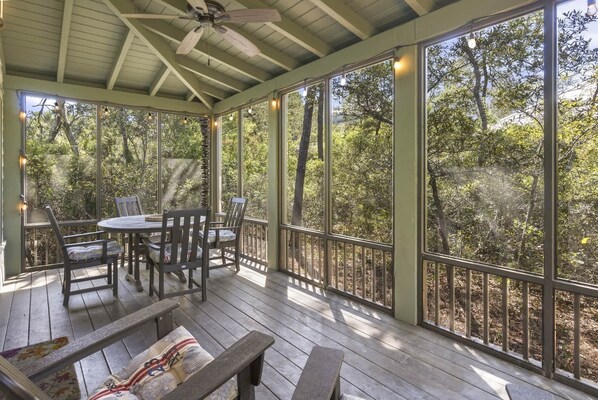A covered outdoor area with a table and chairs, surrounded by large windows offering a view of trees.