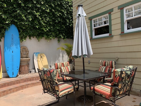 An outdoor patio with a round table and four striped chairs, surrounded by a blue surfboard and a stone sculpture.