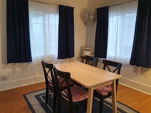 A dining area with a wooden table and four chairs, each with red and white checkered cushions. There are two windows with dark blue curtains, and a wall-mounted fan is visible.