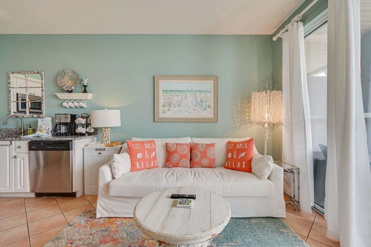 A living room with a white sofa, a round coffee table, a kitchen area with a dishwasher, and a large window with curtains.