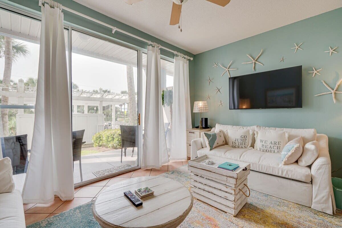 A living room with a sofa, a coffee table, and a TV. There are curtains, a ceiling fan, and a wall with starfish decorations.