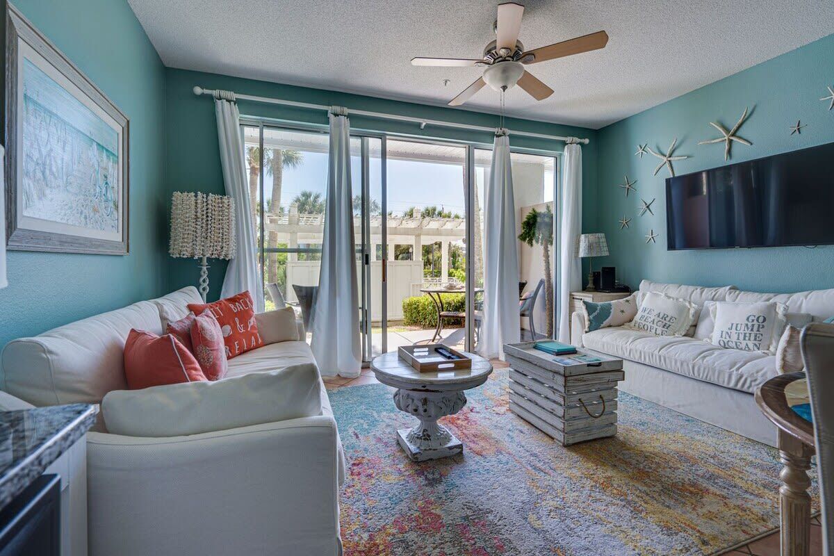 A living room with a white sofa, a coffee table, and a television. There is a ceiling fan and a window with curtains.