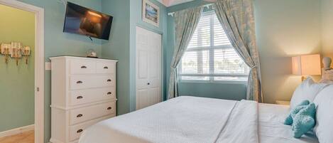 A bedroom with a bed, a ceiling fan, a television, a dresser, and a window with curtains.
