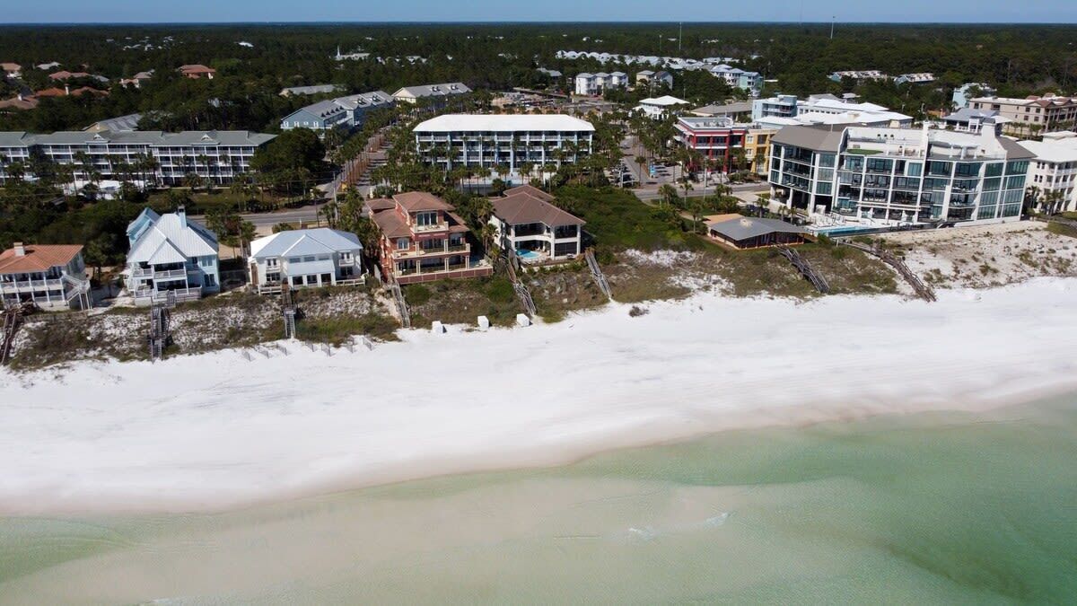 An aerial view of a coastal area with buildings, including a large white structure and several smaller houses.