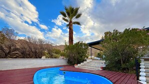 A swimming pool with a wooden deck, surrounded by greenery and a palm tree. - Mid Century Modern - Direct Park Access with Stargazing Patio (Joshua Tree)