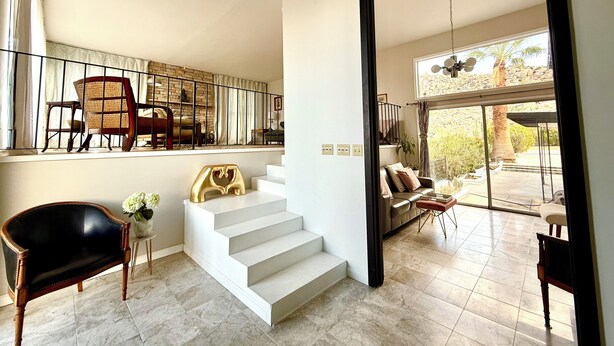 A spacious living area with a staircase leading to an upper level, a balcony with seating, and a view of the outdoors. - Mid Century Modern - Direct Park Access with Stargazing Patio (Joshua Tree)