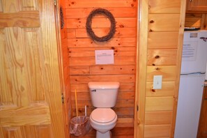 A wooden cabin bathroom with a toilet, a trash bin, and a wreath on the wall. - Blue Rose Cabins - Wildwood Cabin (Logan)