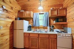 A compact kitchen with wooden walls, a refrigerator, a stove, a microwave, and wooden cabinets. - Blue Rose Cabins - Wildwood Cabin (Logan)