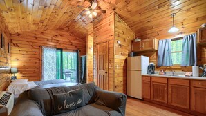 Een knus kamer in chaletstijl met een bed, een bank met kussen, een kitchenette en een raam met gordijnen.