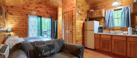 A cozy cabin-style room with a bed, a sofa with a cushion, a kitchenette, and a window with curtains.
