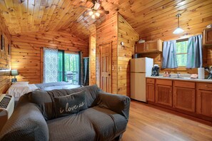 A cozy cabin-style room with a bed, a sofa with a cushion, a kitchenette, and a window with curtains. - Blue Rose Cabins - Wildwood Cabin (Logan)
