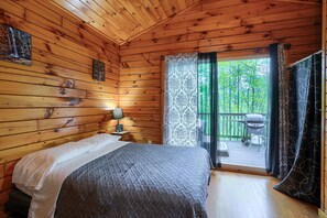 Een slaapkamer in een houten hutje, met een bed, nachtkastje en uitzicht op een terras en bomen.