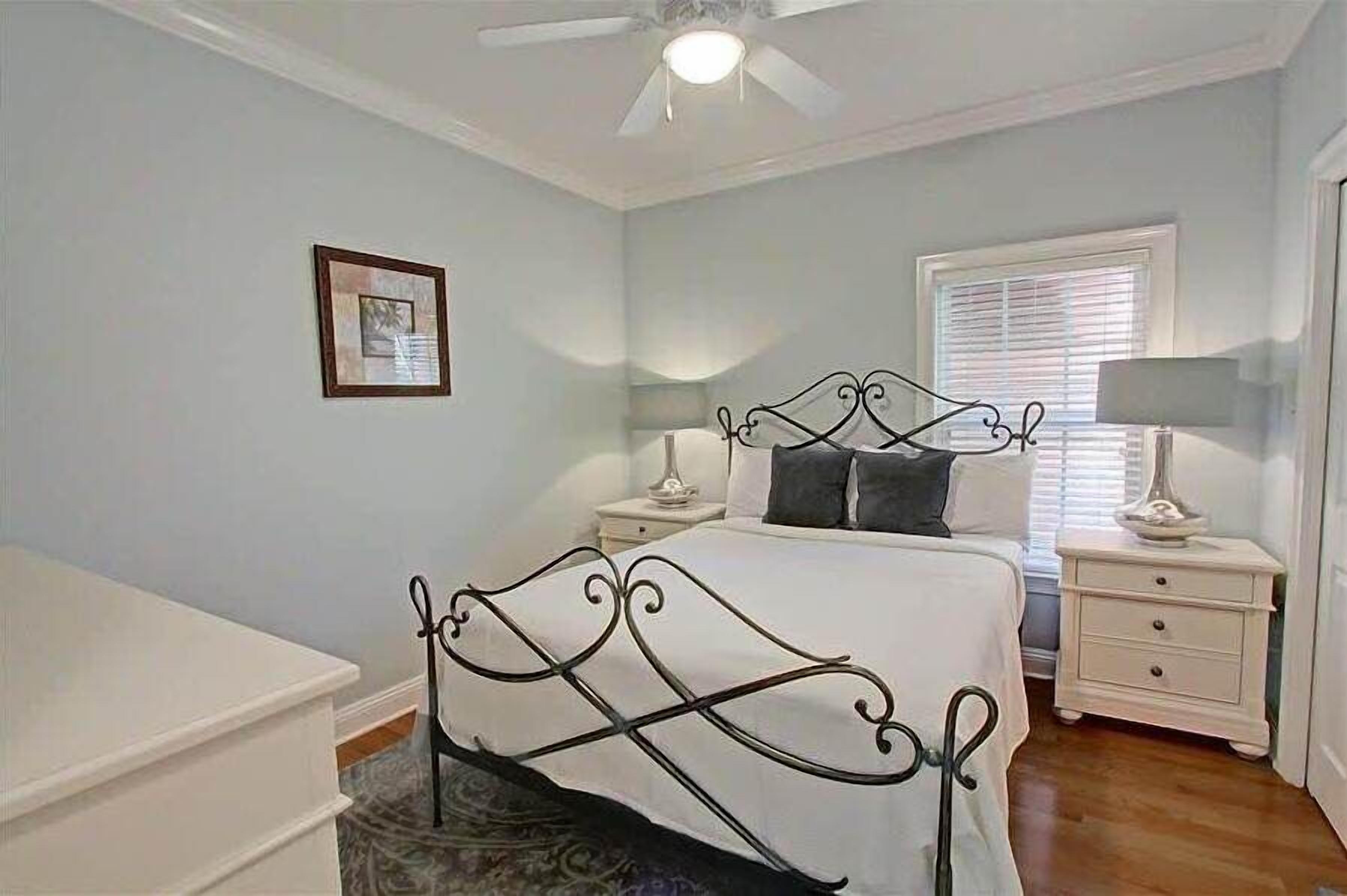 A bedroom with a bed, nightstands, a ceiling fan, a window with blinds, and a framed picture on the wall.