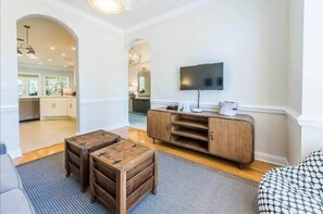 A living room with a wooden coffee table, a TV mounted on the wall, and a kitchen visible through an archway.