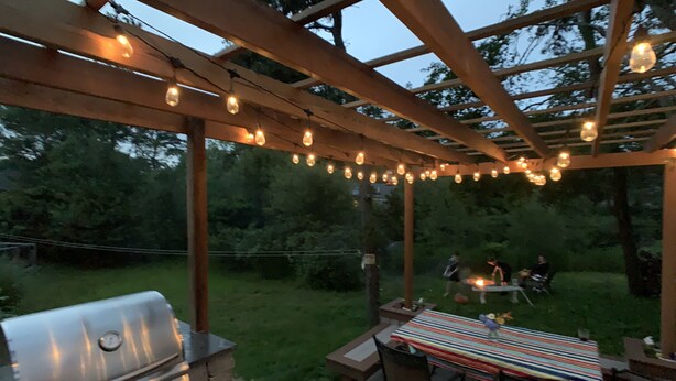 Une terrasse extérieure avec un barbecue, une table recouverte d’une nappe à rayures et des guirlandes lumineuses.