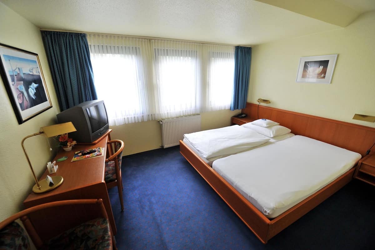 Ein Hotelzimmer mit Bett, Schreibtisch mit Lampe, Fernseher und einem Fenster mit Vorhängen.
