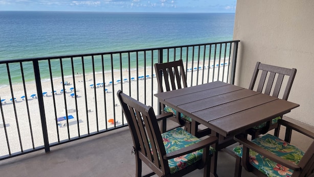 A balcony with a table and chairs, overlooking a beach with umbrellas. - Gulf-Front Phoenix X 3BR  Stunning Balcony Views,  (Orange Beach)