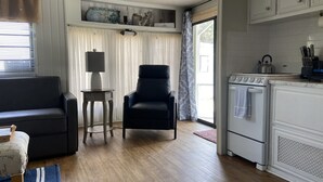 Ein gemütlicher, kleiner Wohnraum mit einer Couch, einem Sessel, einem Tisch mit Lampe, einer Küchenzeile mit Herd und einem Fenster mit Vorhängen.