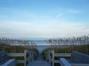 Un paseo marítimo de madera que conduce a una playa con pastos altos y un cielo despejado.