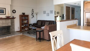 Ett vardagsrum med en öppen spis, en bokhylla, en brun soffa, ett soffbord och en matplats med ett bord och stolar.