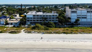 A beachfront hotel with multiple floors, a sandy beach in the foreground, and a view of the sea and distant buildings.