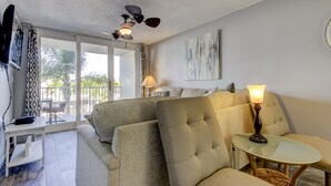 A living room with a sofa, a glass table, a ceiling fan, and a TV mounted on the wall.