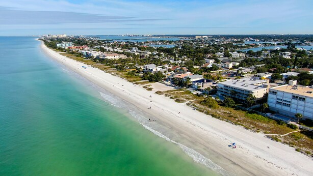 An aerial view of a coastal town with a long sandy beach, residential buildings, and a clear view of the ocean.