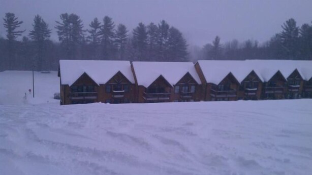 Une rangée de chalets en rondins, dont les toits sont recouverts de neige, s’étend au milieu d’un paysage enneigé et d’une forêt dense.