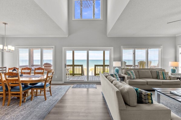 A spacious living room with a large window overlooking the beach, a dining area with wooden furniture, and a cozy seating area with a sofa and cushions. - Namaste at the Beach Oceanfront House, Dog Friendly, Private Beach Access! (Kure Beach)