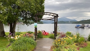 A wooden pathway leading to a wooden archway with a sign, surrounded by colorful flowers and a lake with boats in the background.