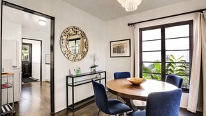 A dining area with a round table, blue chairs, a chandelier, and a large window with curtains.
