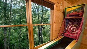 Una habitación con un ventanal grande que da a un bosque, techo de madera y una máquina de skee-ball.