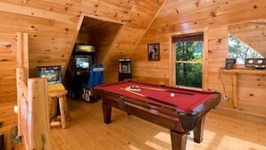 Una sala de madera con mesa de billar, juegos de arcade y un ventanal grande.