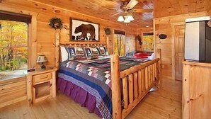Dormitorio de cabaña de madera con cama, mesita de noche, ventilador de techo y ventana con vistas a árboles otoñales.
