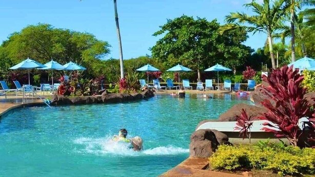 A swimming pool with lounge chairs and umbrellas, surrounded by palm trees and tropical plants. - Oceanview, Walk to Poipu Beach, AC, Private Lanai! (Koloa)