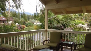 A covered outdoor seating area with wicker furniture and a glass table, surrounded by lush greenery and a view of distant houses. - Magical 4 Bedroom 3 Bath Home with Awesome Mountain View's. New Full house AC (Princeville)